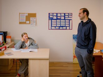 Eine Frau sitzt an einem Schreibtisch und stützt ihr Kinn auf die Hand, während sie zu einem lächelnden Mann aufblickt, der neben ihr steht. Der Mann trägt eine Brille und eine dunkelblaue Jacke. Im Hintergrund hängen ein Korkbrett und eine blaue Planungsübersicht an der Wand.