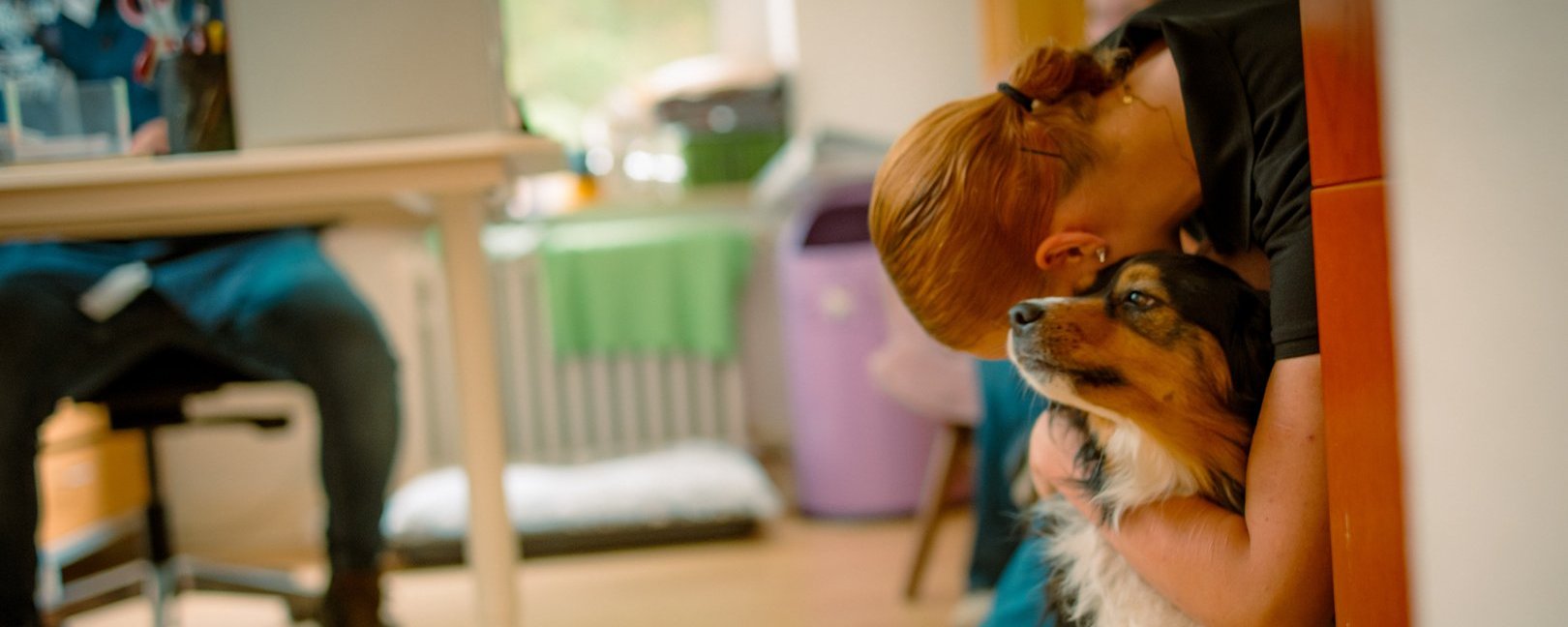 Eine Frau mit roten Haaren lehnt an einer Wand und umarmt zärtlich einen braun-weißen Hund, der zu ihr aufblickt. Im unscharfen Hintergrund ist ein Büroarbeitsplatz zu erkennen.