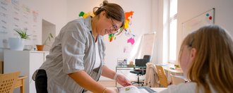 Eine Frau mit Brille steht an einem Tisch und erklärt einer sitzenden Schülerin etwas anhand eines aufgeschlagenen Buches. Im Hintergrund ist ein heller Raum mit bunten Wanddekorationen zu sehen.