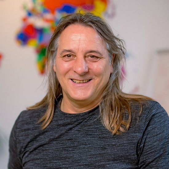 Ein Mann mit langen Haaren und einem grauen T-Shirt lächelt in die Kamera. Im unscharfen Hintergrund ist eine bunte Weltkarte an der Wand zu erkennen.