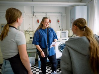 Drei junge Frauen unterhalten sich in einem Waschraum. Die Frau in der Mitte hält ein Blatt Papier und scheint Anweisungen zu geben, während die anderen beiden ihr zuhören. Im Hintergrund sind Waschmaschinen zu sehen.