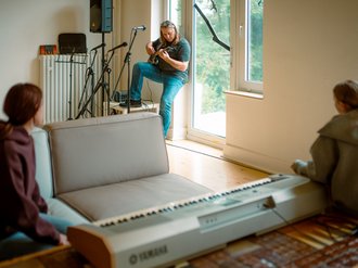 Ein Mann spielt E-Gitarre in einem hellen Raum vor einem Fenster. Zwei Personen sitzen im Vordergrund und beobachten sie. Auf einem Tisch steht ein weißes Keyboard.