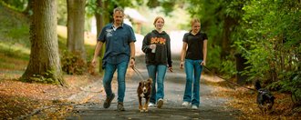 Ein Mann und zwei Frauen gehen mit zwei Hunden an der Leine einen asphaltierten Weg durch einen grünen, bewaldeten Park entlang.
