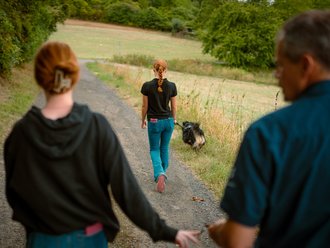 Drei Personen und ein Hund gehen auf einem Kiesweg durch eine grüne Landschaft weg von der Kamera. Im Vordergrund sind zwei Personen teilweise sichtbar, während eine Frau mit roten Haaren und ein Hund weiter vorne auf dem Pfad gehen.