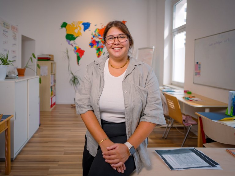 Eine lächelnde Frau mit Brille steht in einem Klassenzimmer. Sie trägt ein hellgraues Hemd. Im Hintergrund hängt eine bunte Weltkarte. Links steht ein Schrank mit Pflanzen, rechts sind Tische und ein Fenster zu sehen.