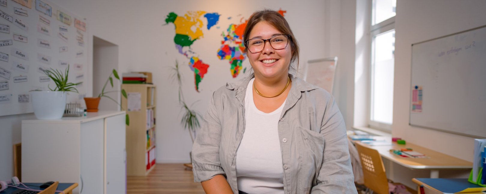 Eine lächelnde Frau mit Brille steht in einem Klassenzimmer. Sie trägt ein hellgraues Hemd. Im Hintergrund hängt eine bunte Weltkarte. Links steht ein Schrank mit Pflanzen, rechts sind Tische und ein Fenster zu sehen.