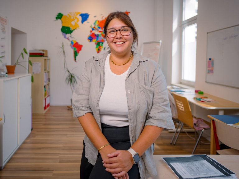 Eine lächelnde Frau mit Brille steht in einem Klassenzimmer. Sie trägt ein hellgraues Hemd. Im Hintergrund hängt eine bunte Weltkarte. Links steht ein Schrank mit Pflanzen, rechts sind Tische und ein Fenster zu sehen.